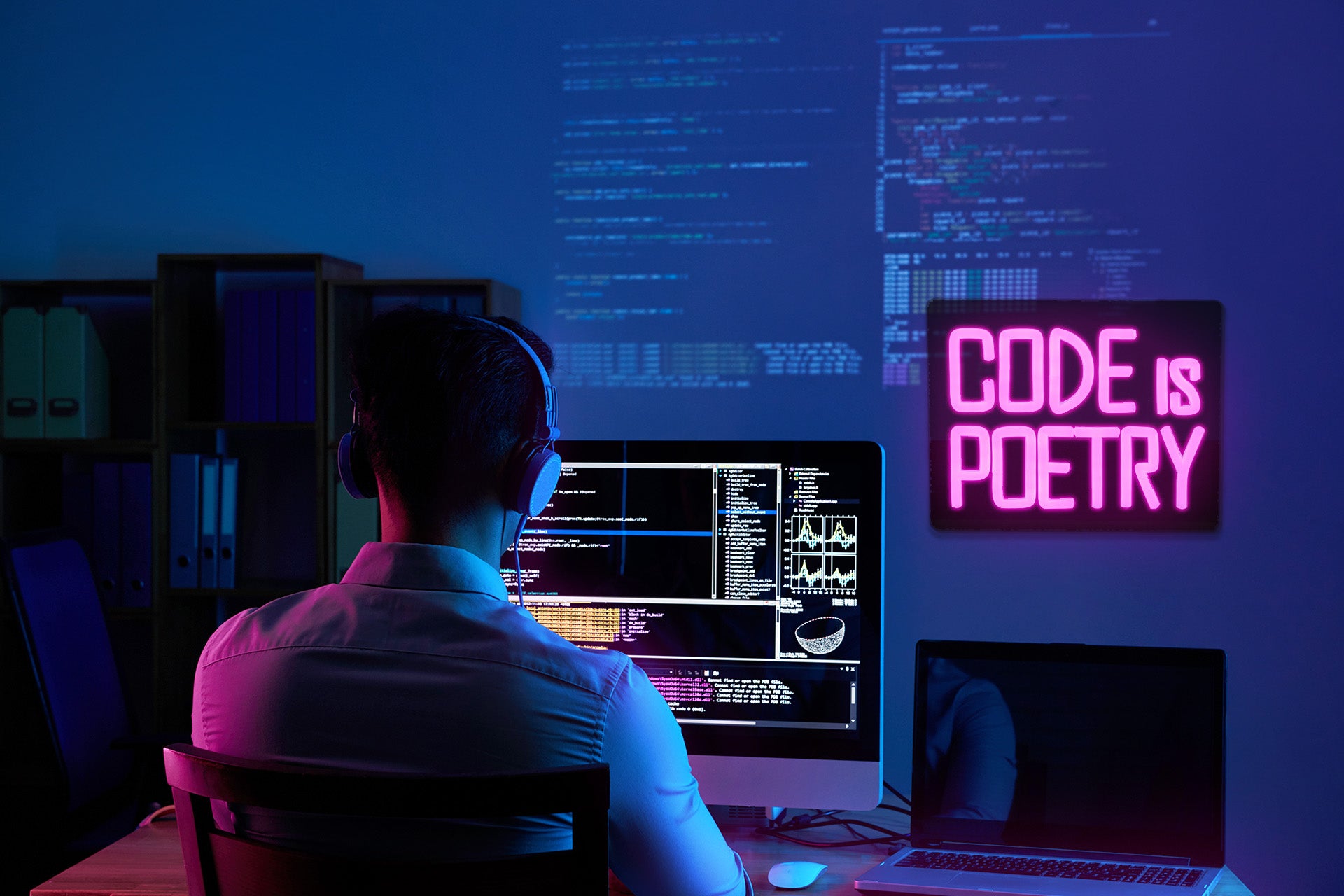 A developer sits in front of a computer with a neon sign on the wall that reads "Code is Poetry"