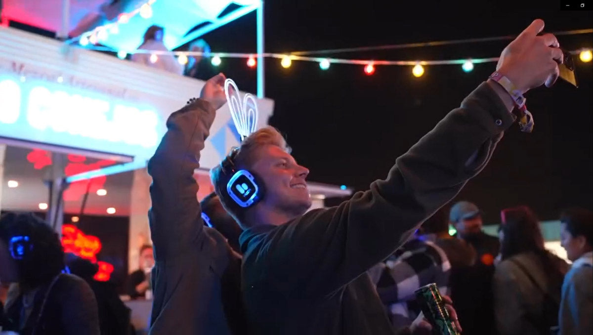 Man taking a selfie at a nighttime event with colorful lights and people in the background